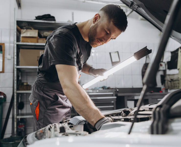 Car service, repair, maintenance and people concept - car mechanic man with lamp working at workshop