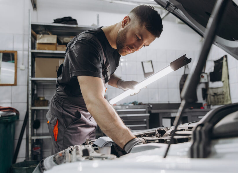 Car service, repair, maintenance and people concept - car mechanic man with lamp working at workshop