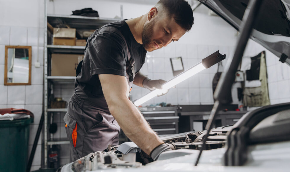 Car service, repair, maintenance and people concept - car mechanic man with lamp working at workshop